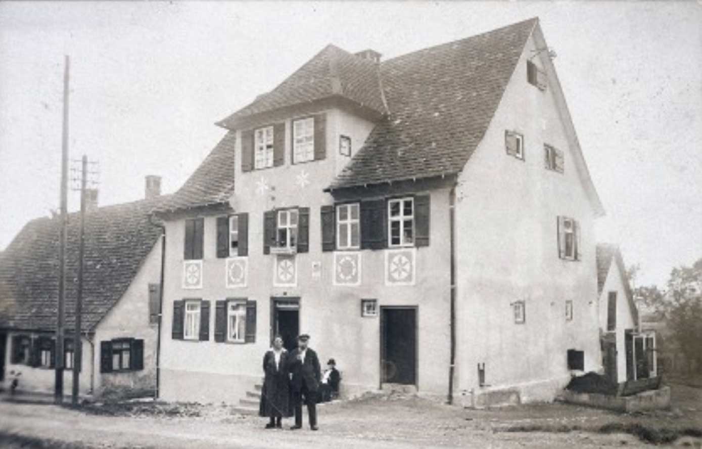 Schwarz-Weiß-Foto eines historischen Hauses mit zwei Personen und einem Kind im Vordergrund, umgeben von ländlicher Idylle.