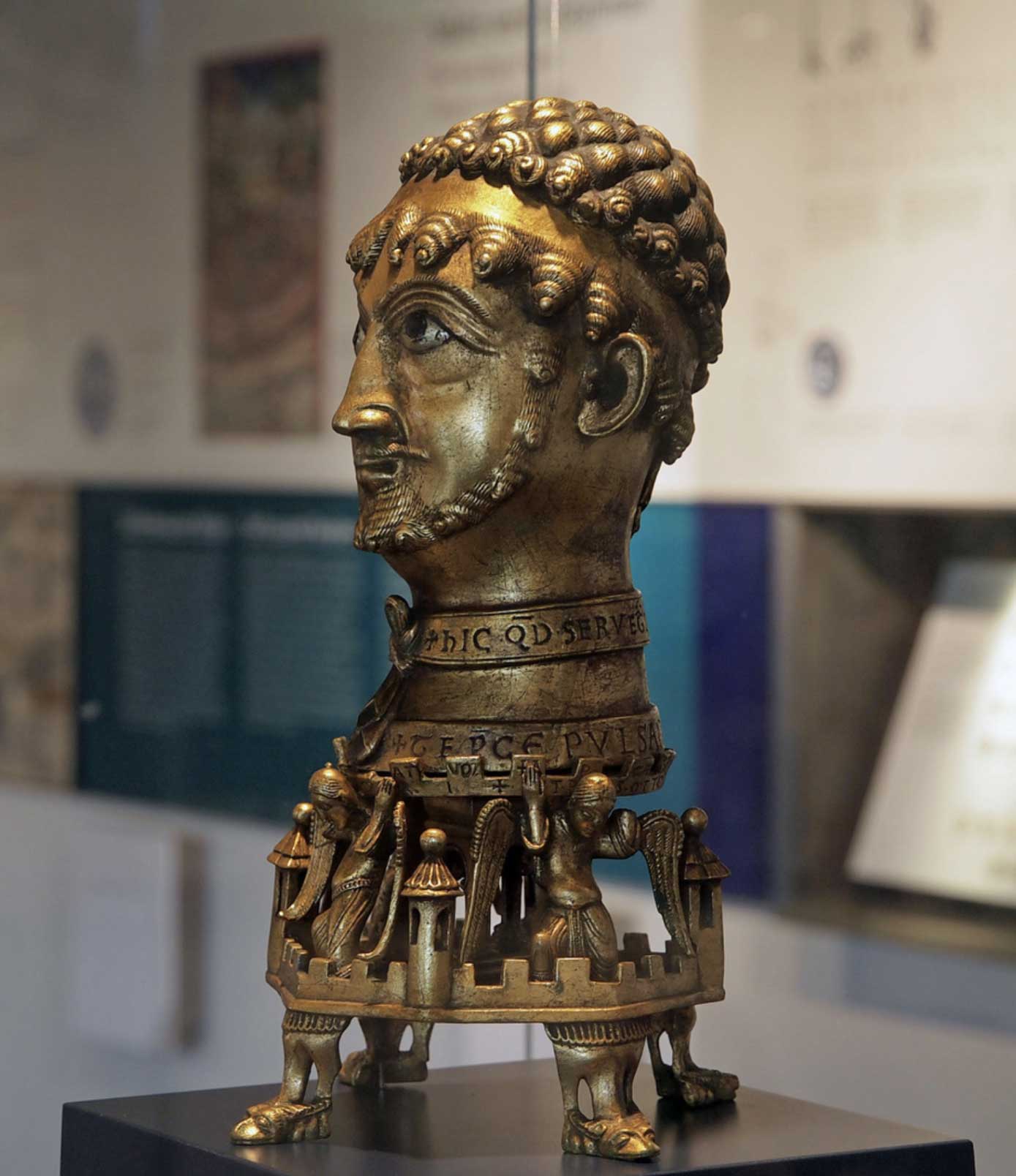 Bronzeskulptur eines Kopfes mit detaillierten Verzierungen und Figuren am Sockel, in einem Museum ausgestellt.