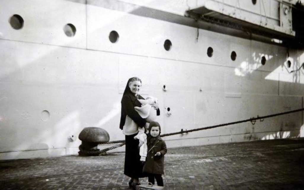 Eine Frau hält ein Baby, während ein kleines Kind neben ihr steht, vor einem großen Schiff.