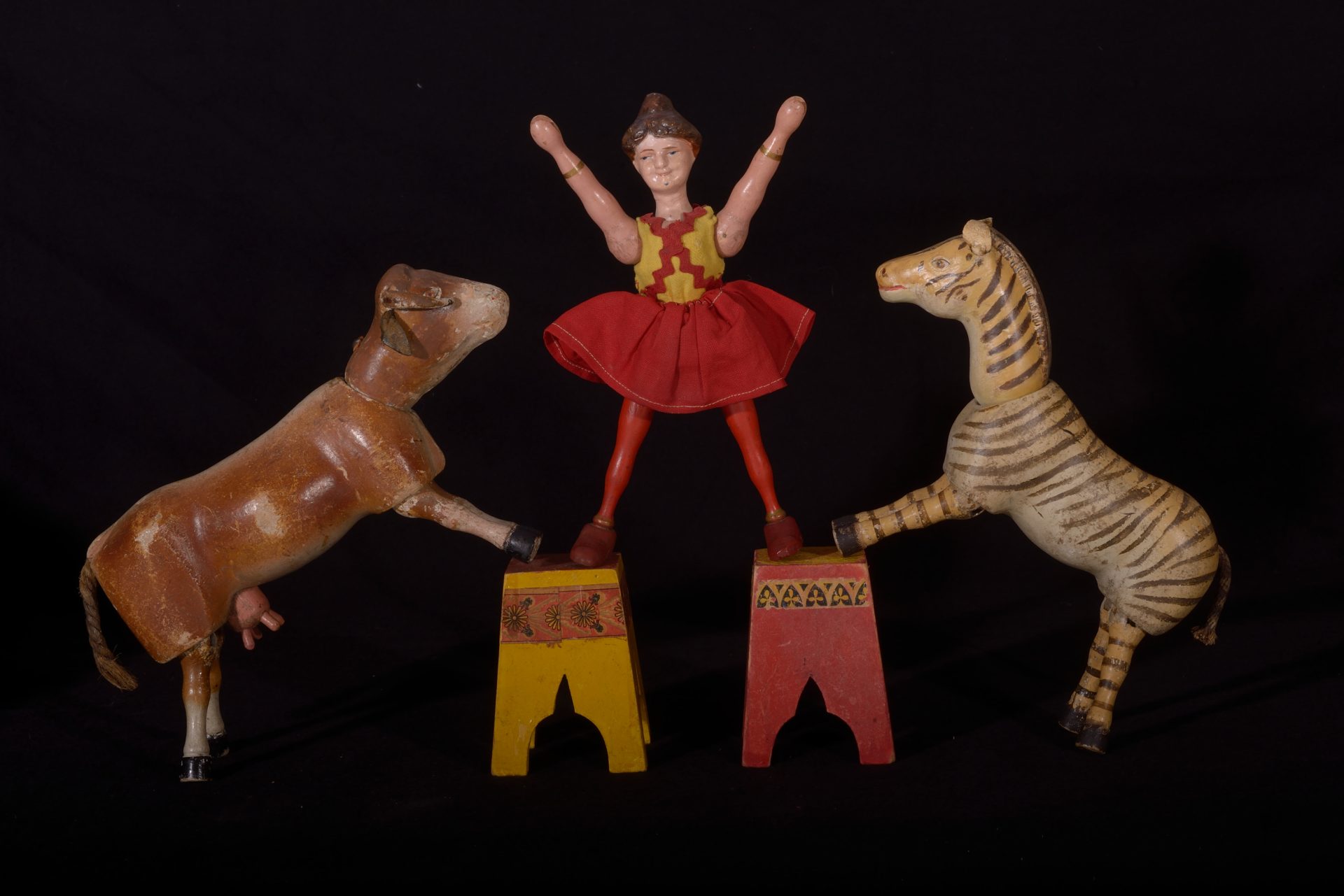 Zu sehen sind Gliederfiguren aus Holz. Es handelt sich um eine Artistin, eine Kuh und ein Zebra sowie zwei Hocker. Die Figuren und die Hocker sind farbig gestaltet, die Artistin ist eingekleidet.