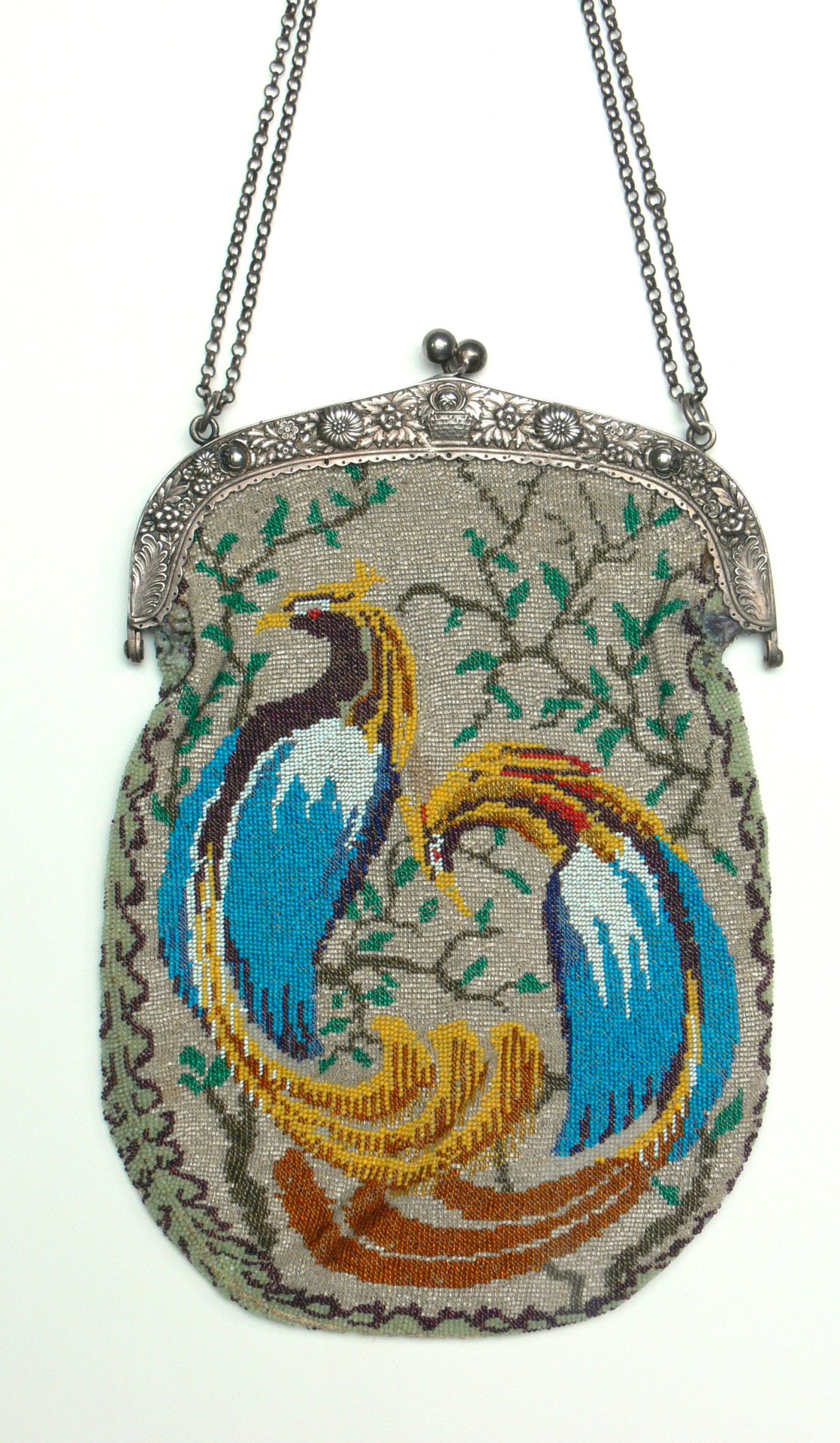 Eine handgestrickte Handtasche aus winzigen farbigen Glasperlen. Dargestellt sind zwei blau-gelbe Vögel mit langen Schwänzen. Die Tasche besitzt einen Silberbügel und eine daran gefestigte Kette.